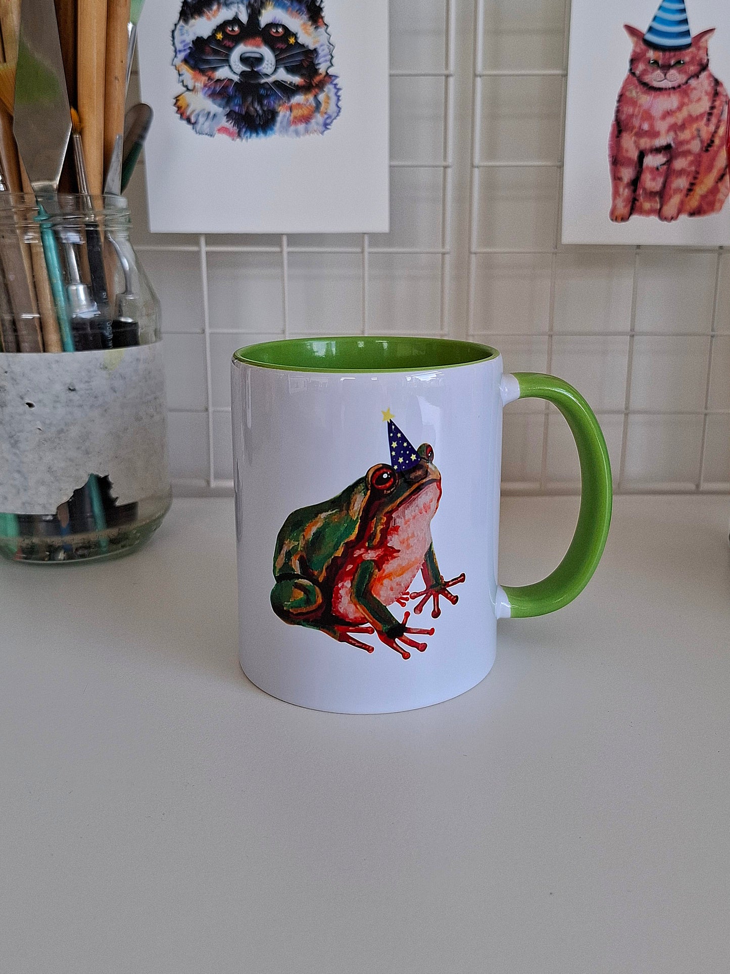 White ceramic mug with green handle and interior featuring a cute green frog wearing a magician hat