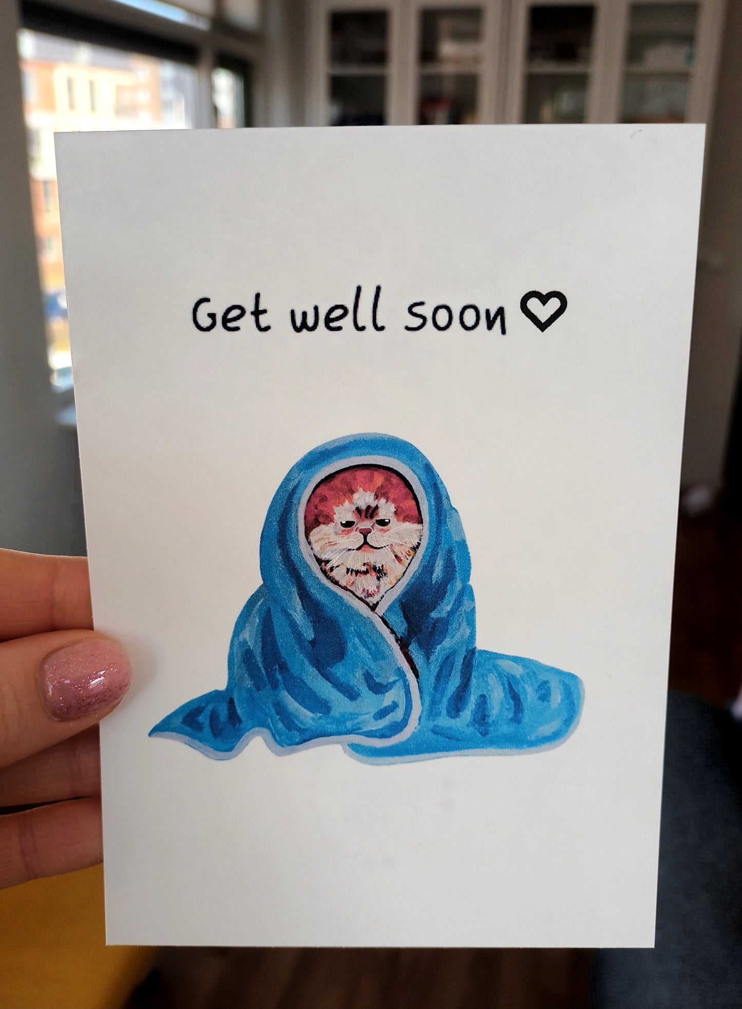 Handmade Get Well Soon greeting card featuring a cute cat wrapped in a cozy blanket, printed on eco-friendly paper held by hand