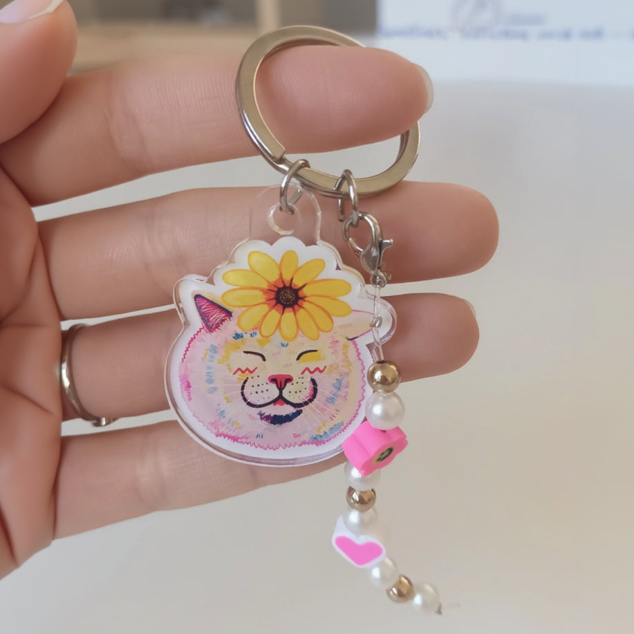 An acrylic keychain featuring a white cat with a flower on its head, accompanied by a stainless steel key ring and decorative elements in pink and yellow.