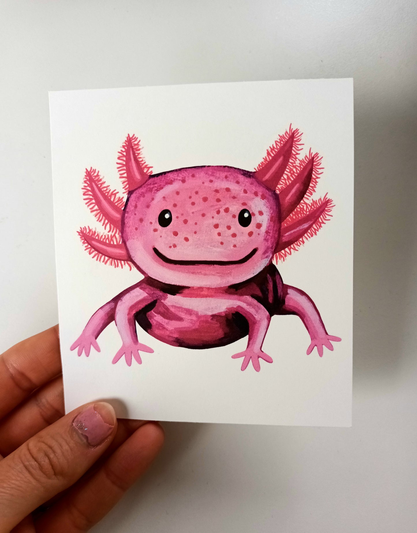 Hand-painted pink axolotl with a happy smile on a soft background, printed on a square eco-friendly postcard held by hand