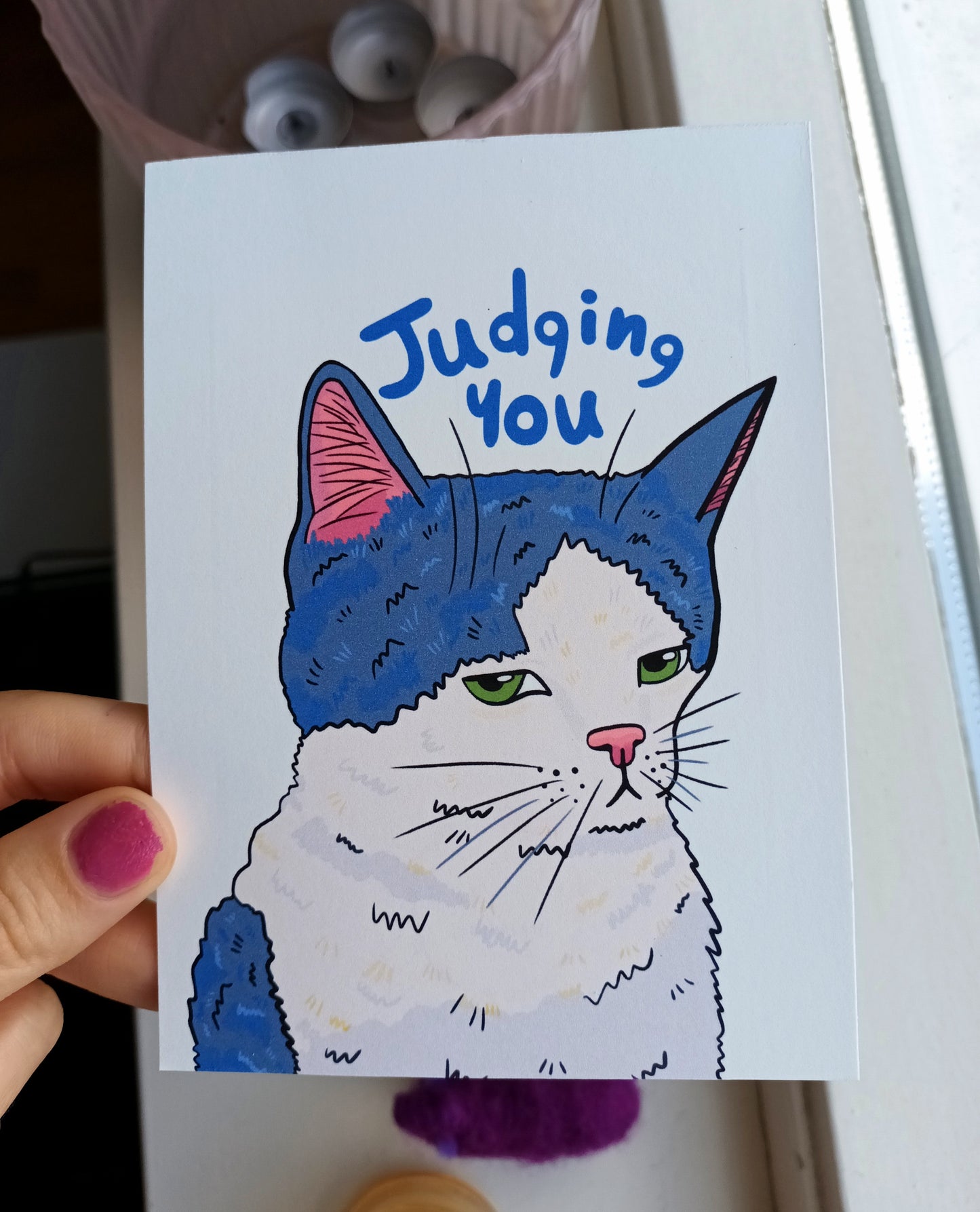 Card with a cat illustration and text 'Judging You' held by a hand.