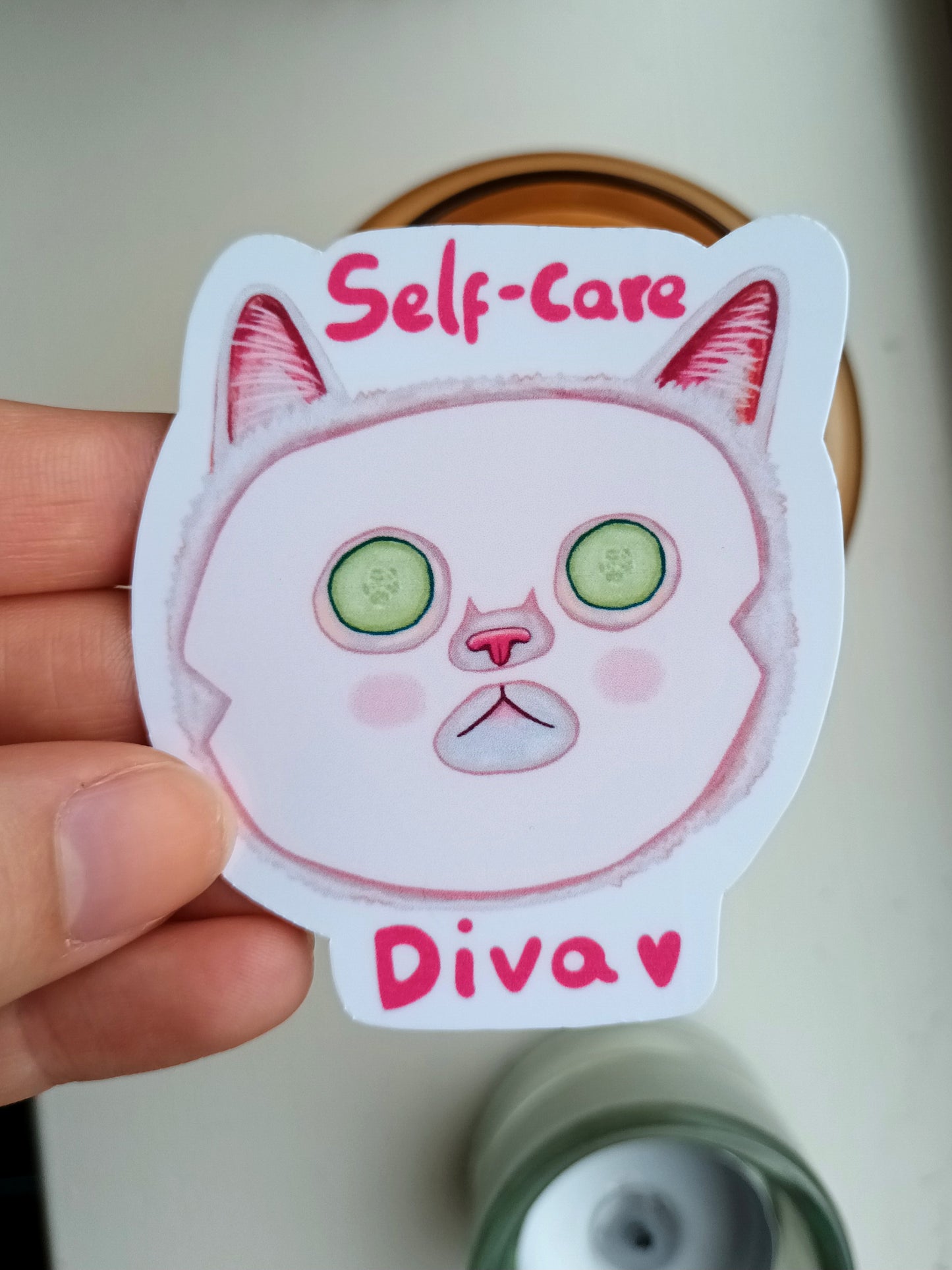 Sticker of a cat face with 'Self-care Diva' text held by a hand.