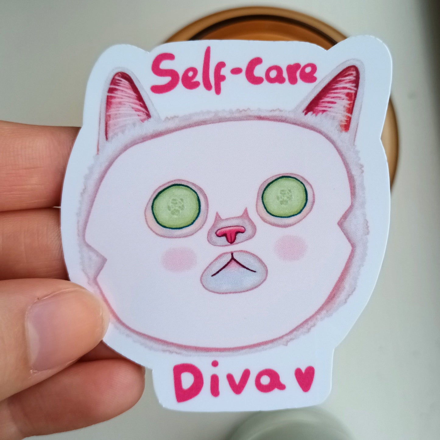 Sticker of a cat face with 'Self-care Diva' text held by a hand.