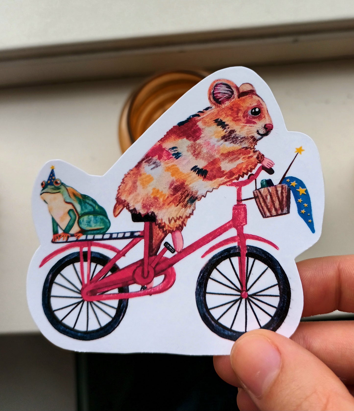Vinyl sticker of a hamster riding a bike with a frog in a magician hat on the back, hand-drawn and colourful.