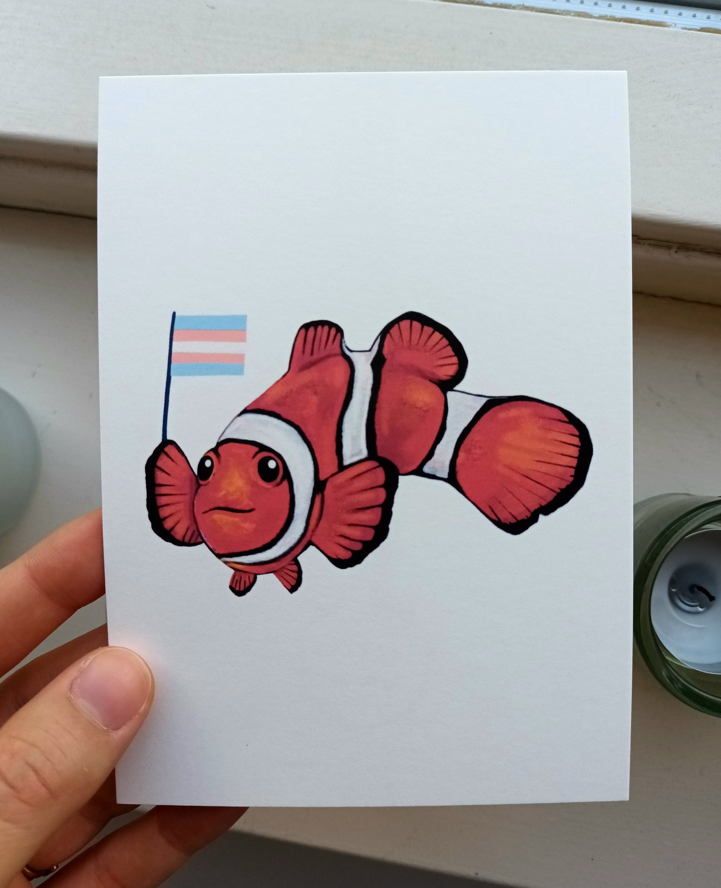 Cute clownfish holding trans flag postcard – handmade LGBTQ+ pride card.