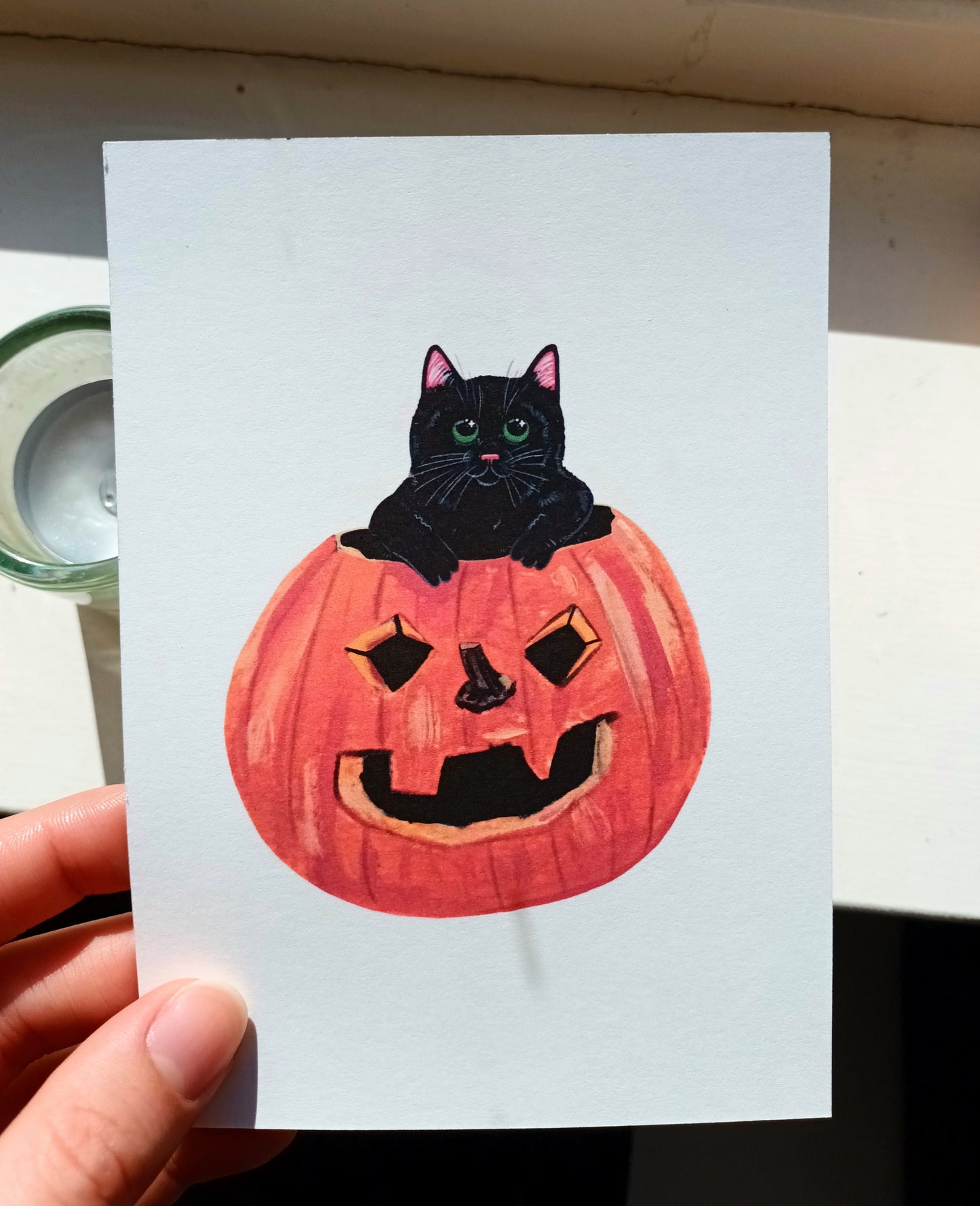 Card with a black cat in a pumpkin, held by a hand.