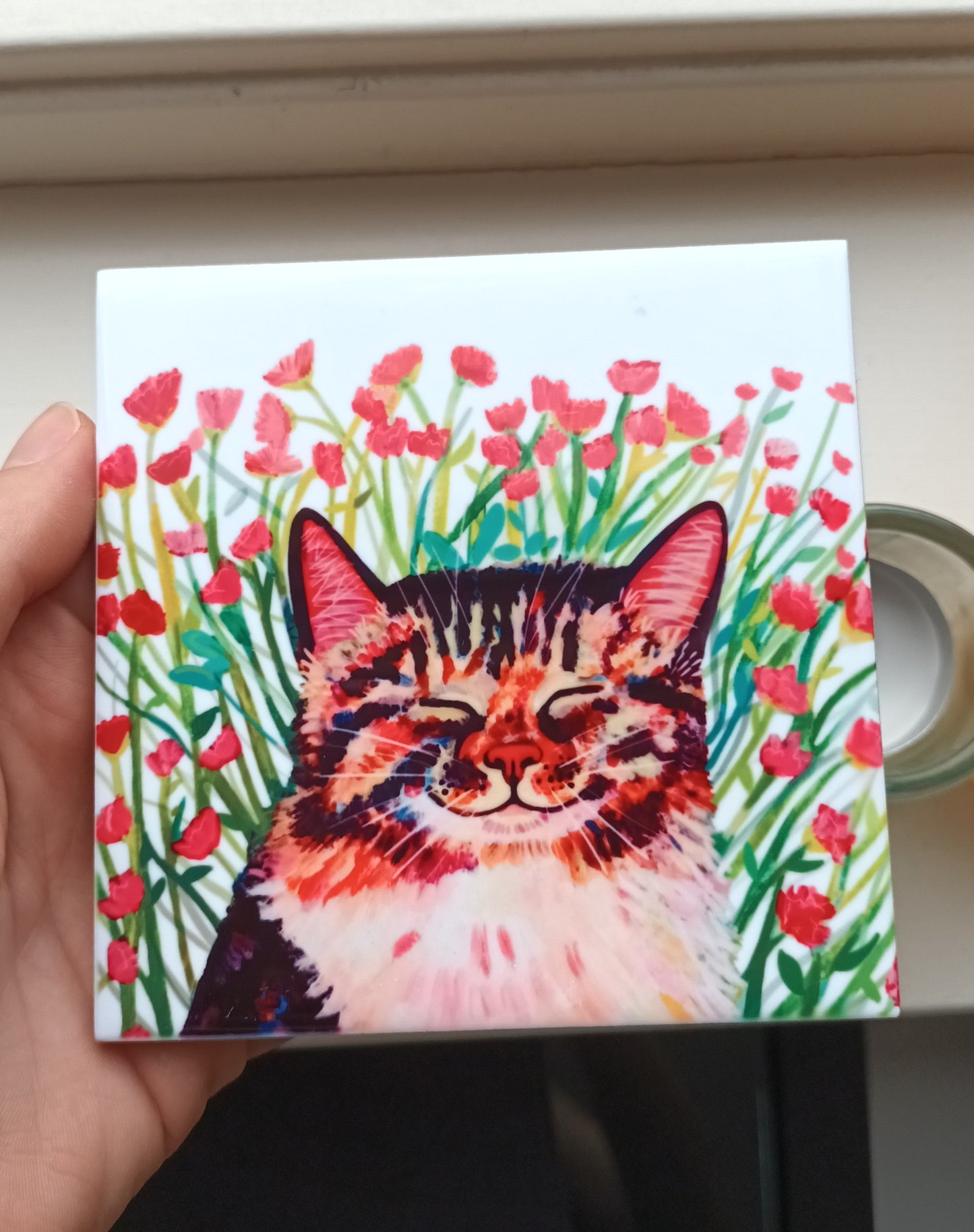 Square tile with a colorful illustration of a cat among flowers, held by a hand.