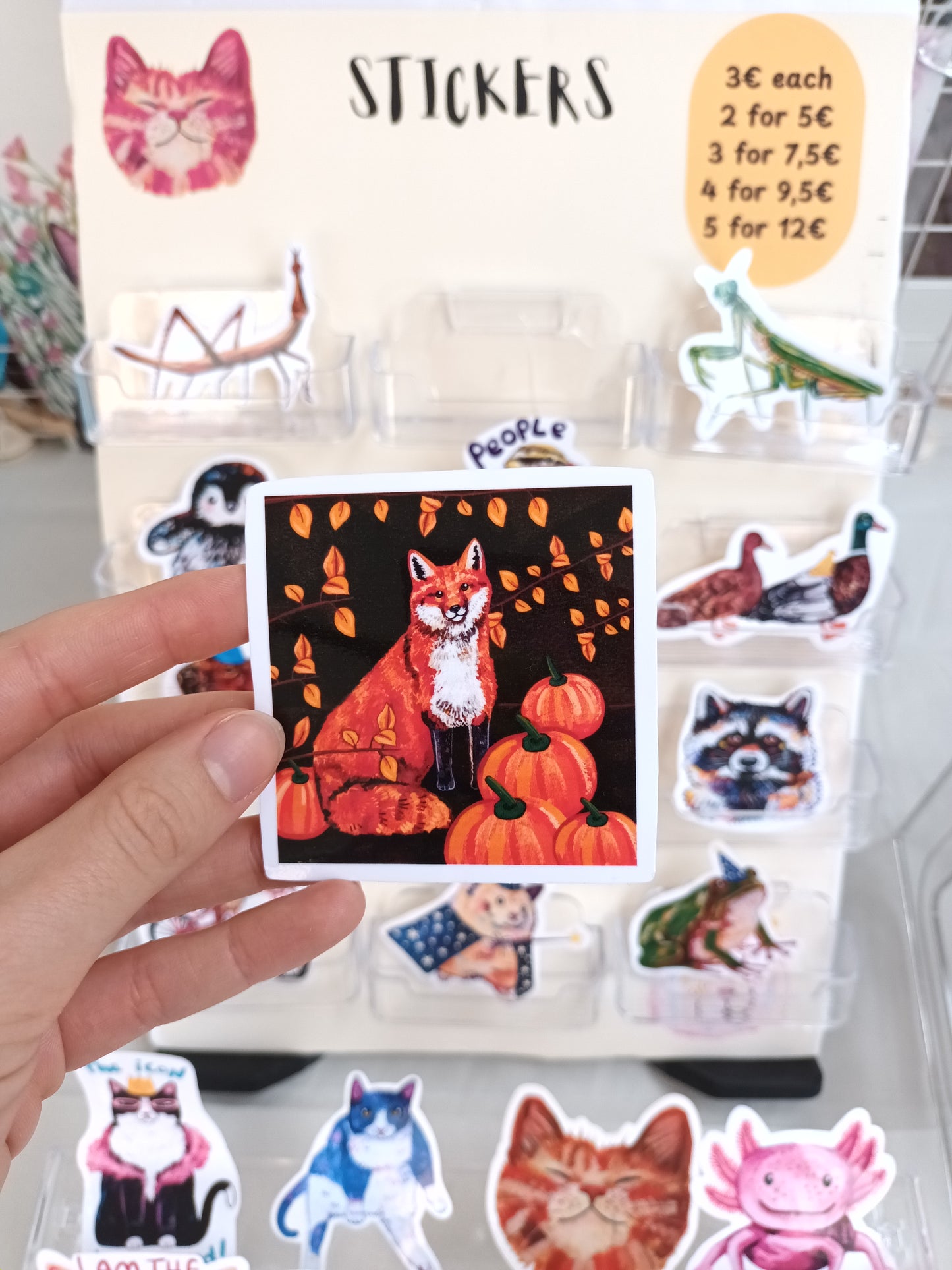 Hand holding a sticker with a fox and pumpkin design, surrounded by other stickers on a white surface.