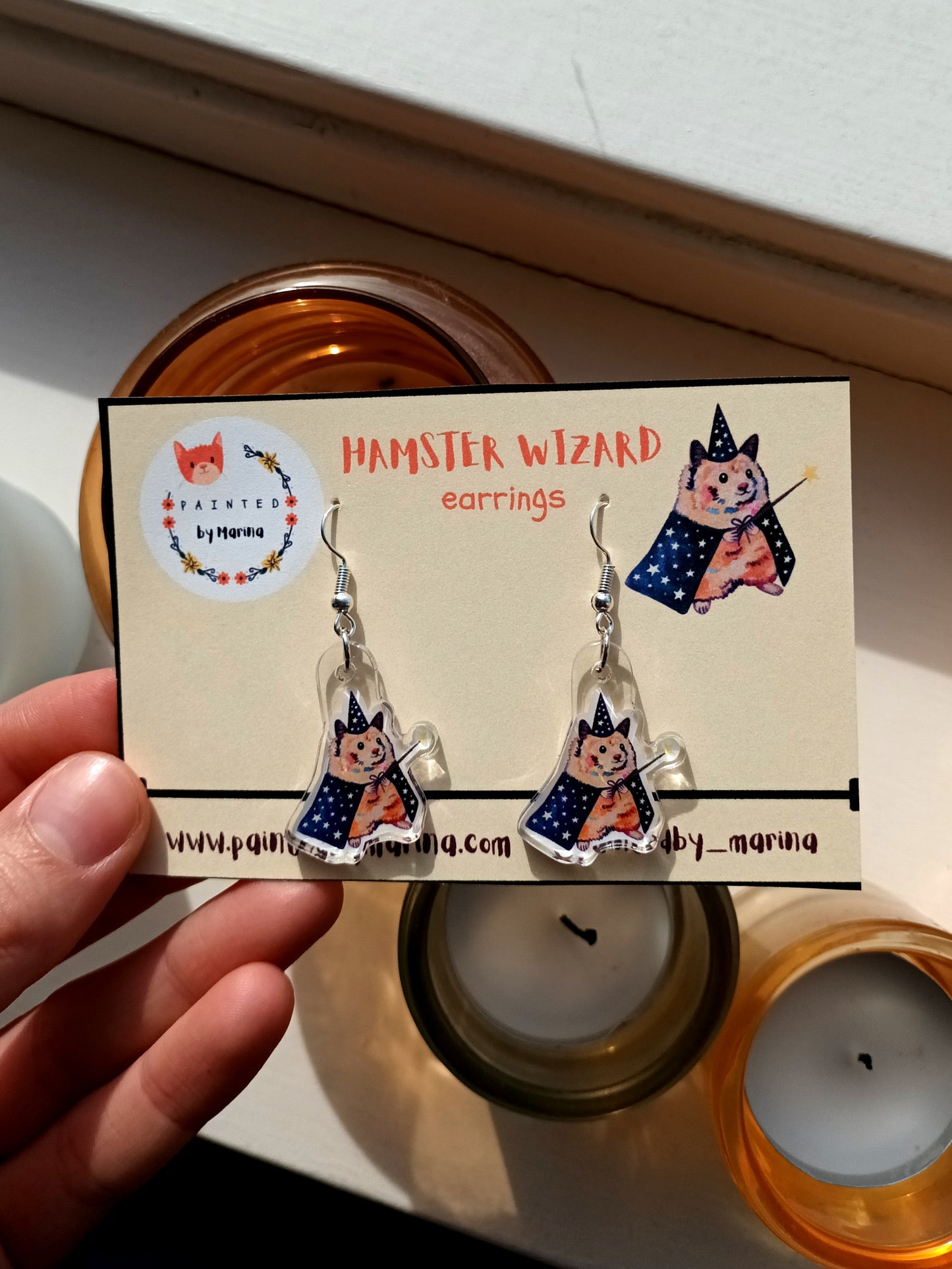 Hamster wizard earrings on a branded card held by a hand with candles in the background.