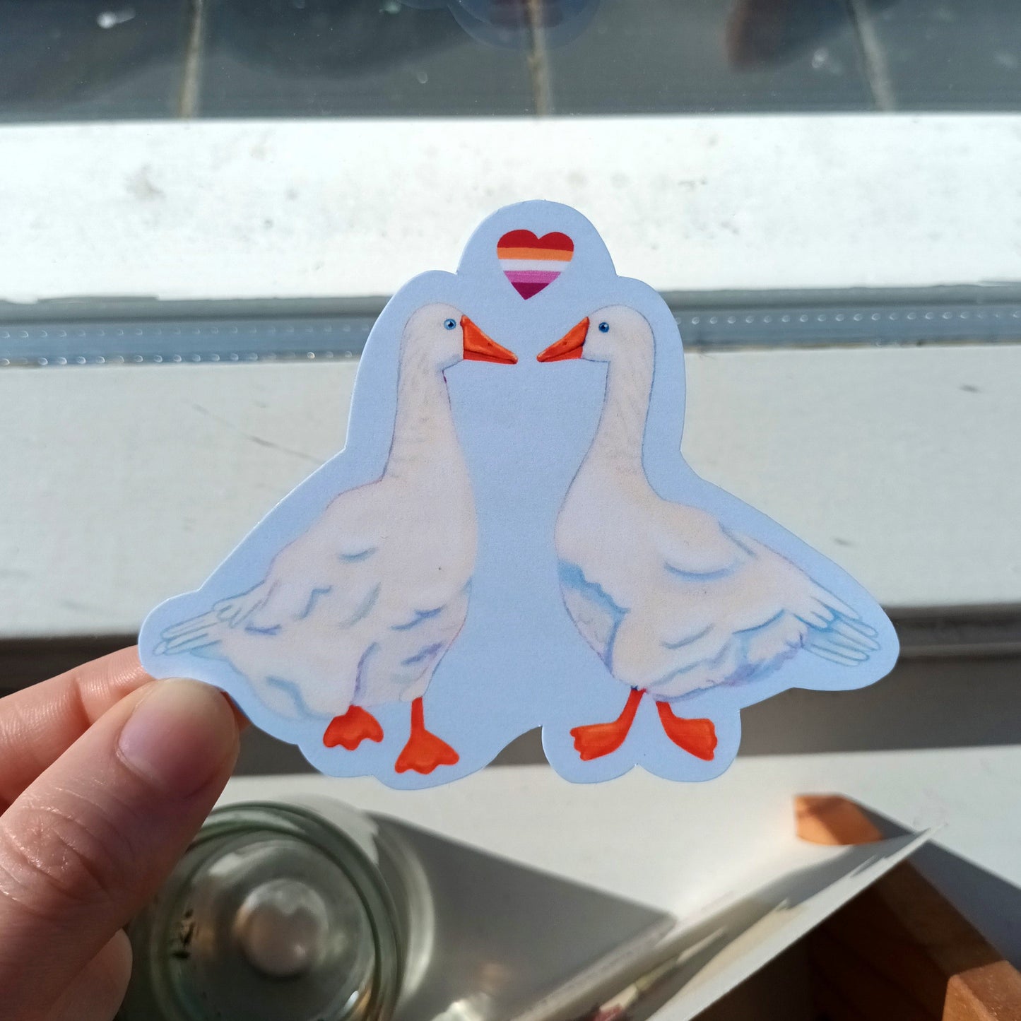 Hand holding a sticker of a cute lesbian geese couple.