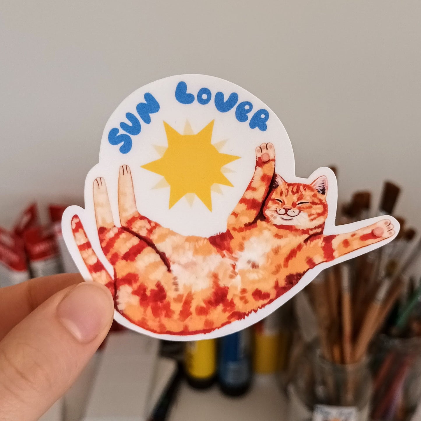 Sticker of a cat labeled 'Sun Lover' held by a hand with art supplies in the background.