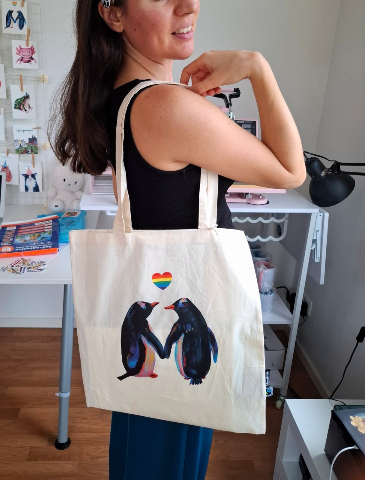 Person holding a tote bag with penguin design and rainbow heart in a room.