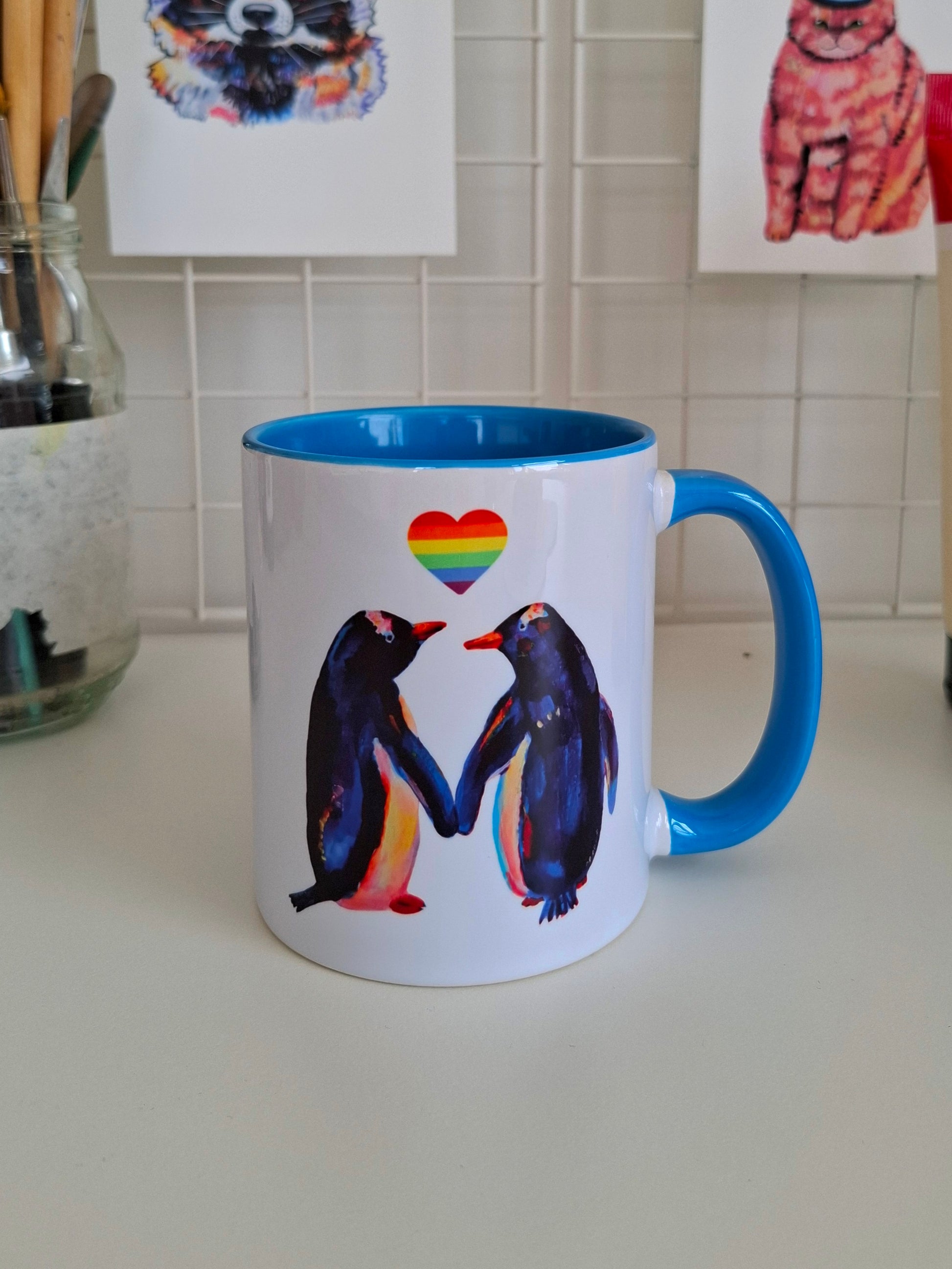 Mug with penguins holding hands and a rainbow heart on a kitchen counter.