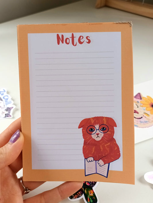 Orange cat studying illustration on an A6 notepad with blank pages and thick cardboard backing, available in 30 or 50 page options