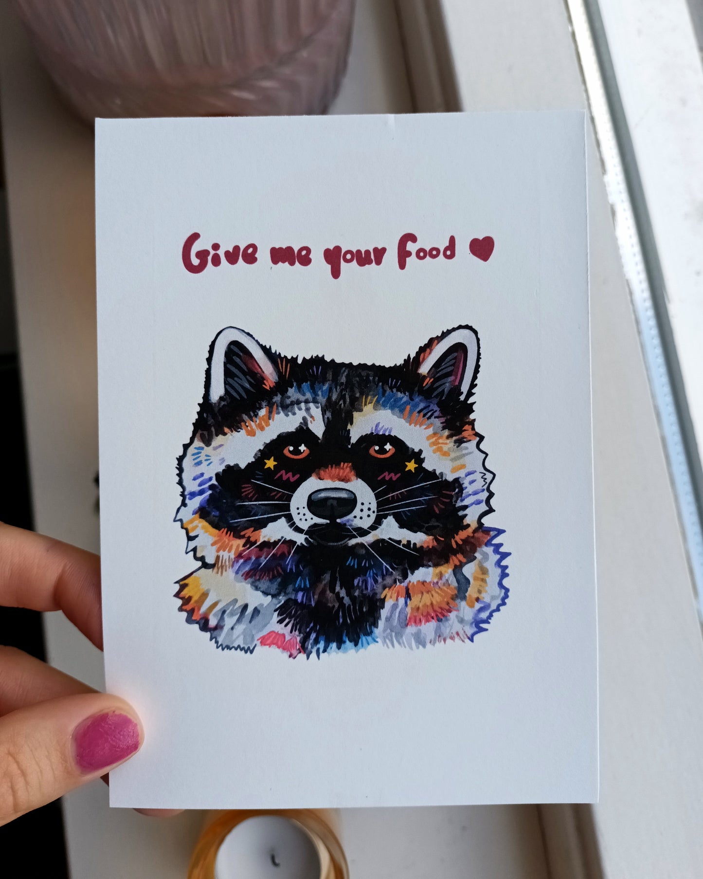 Card with a colorful raccoon illustration and text 'Give me your food' held by a hand.