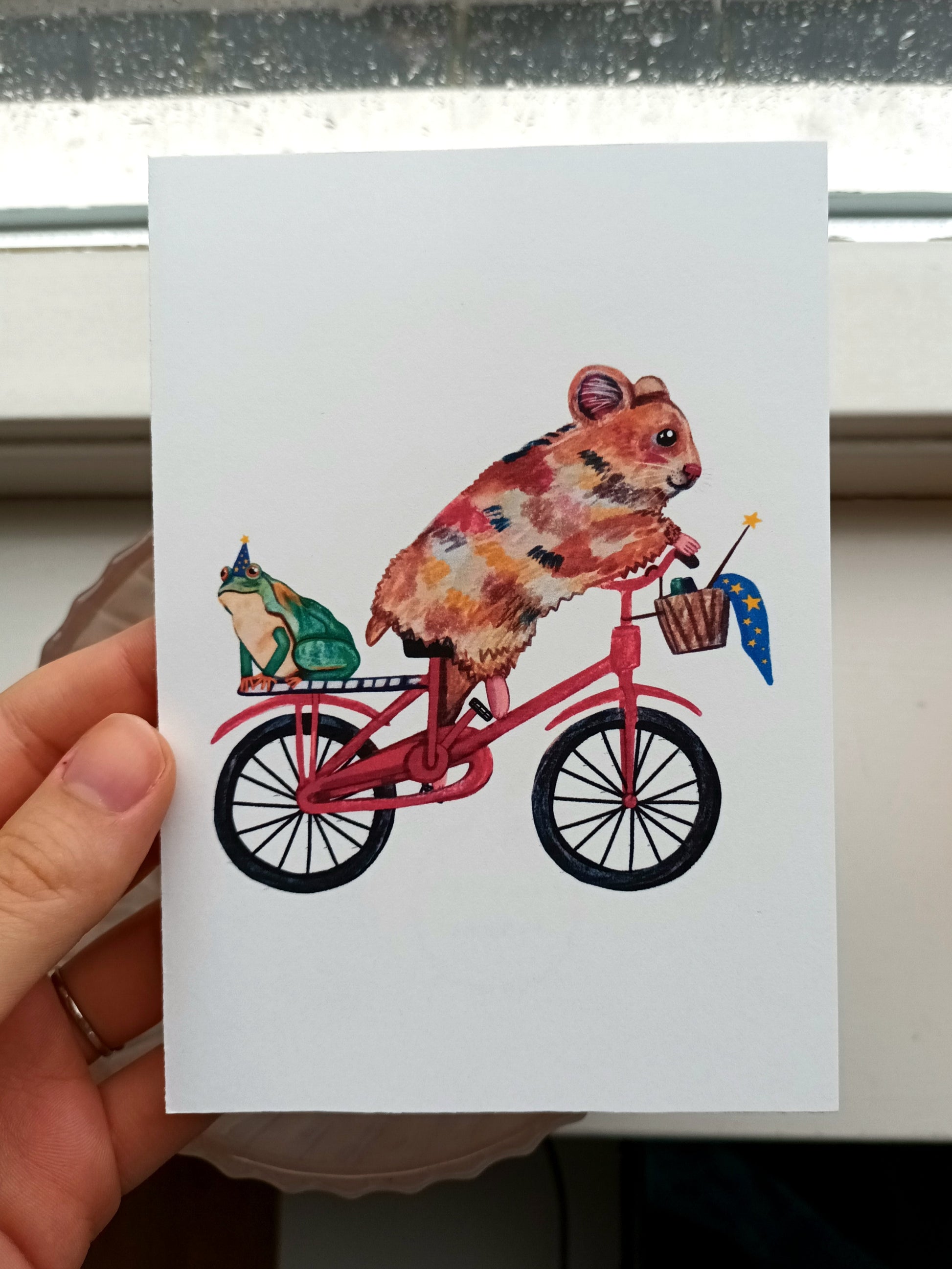 Postcard with a hamster riding a bike and a frog in a magician hat on the back, printed on eco-friendly paper.
