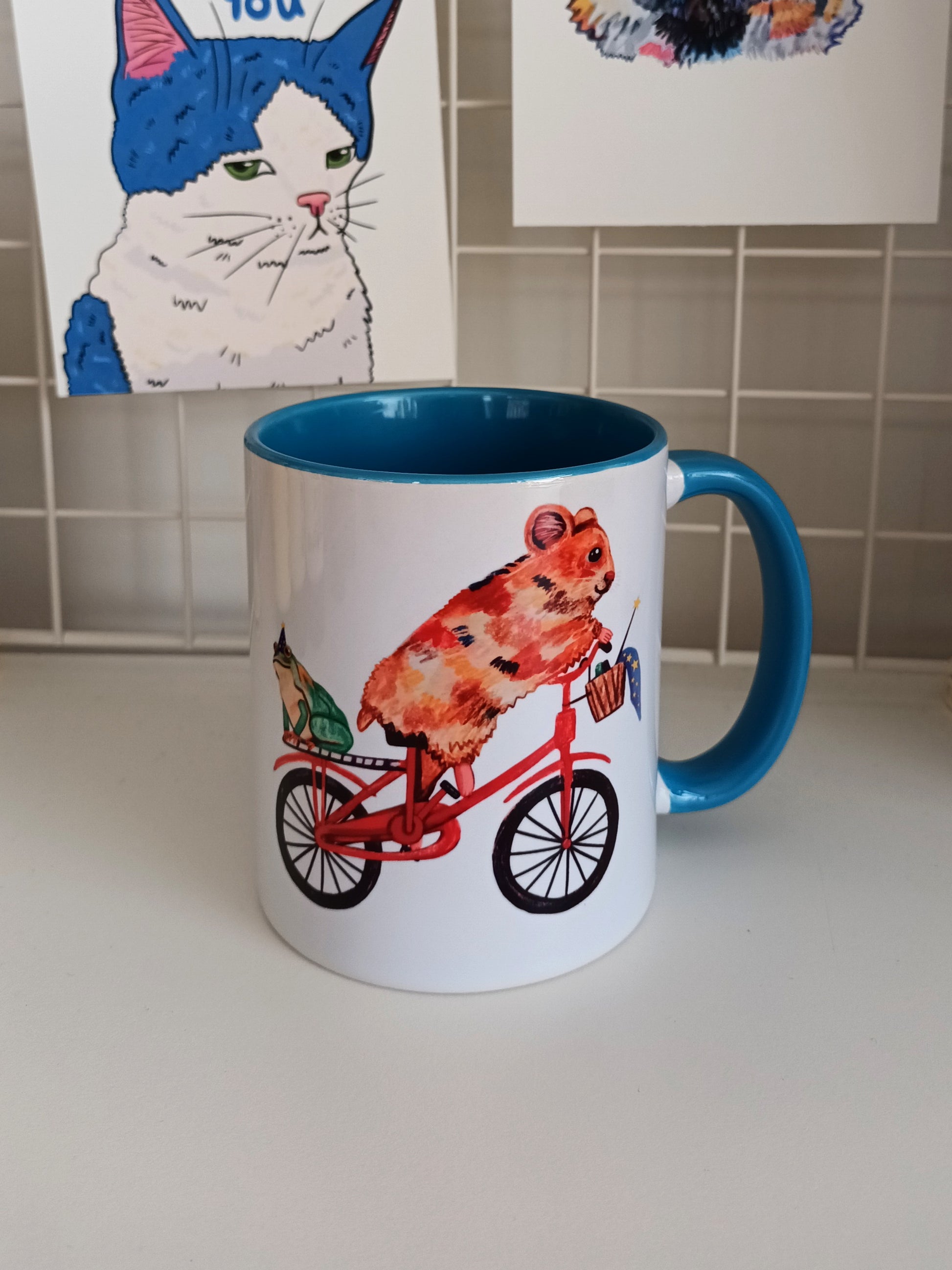 Handmade ceramic mug with a cute hamster riding a bike and a frog in a magician hat on the back, blue handle and interior.