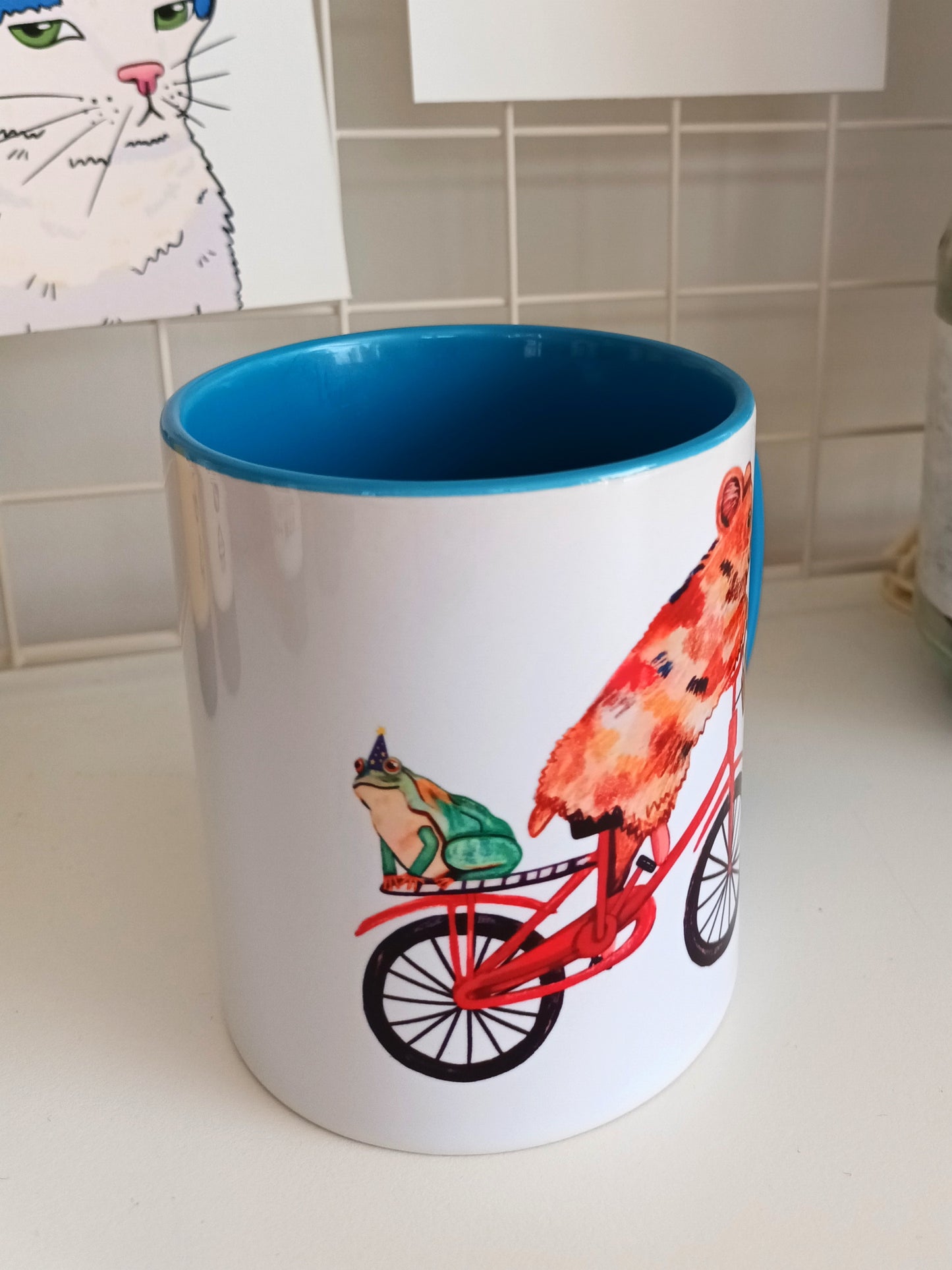 Handmade ceramic mug with a cute hamster riding a bike and a frog in a magician hat on the back, blue handle and interior side view.