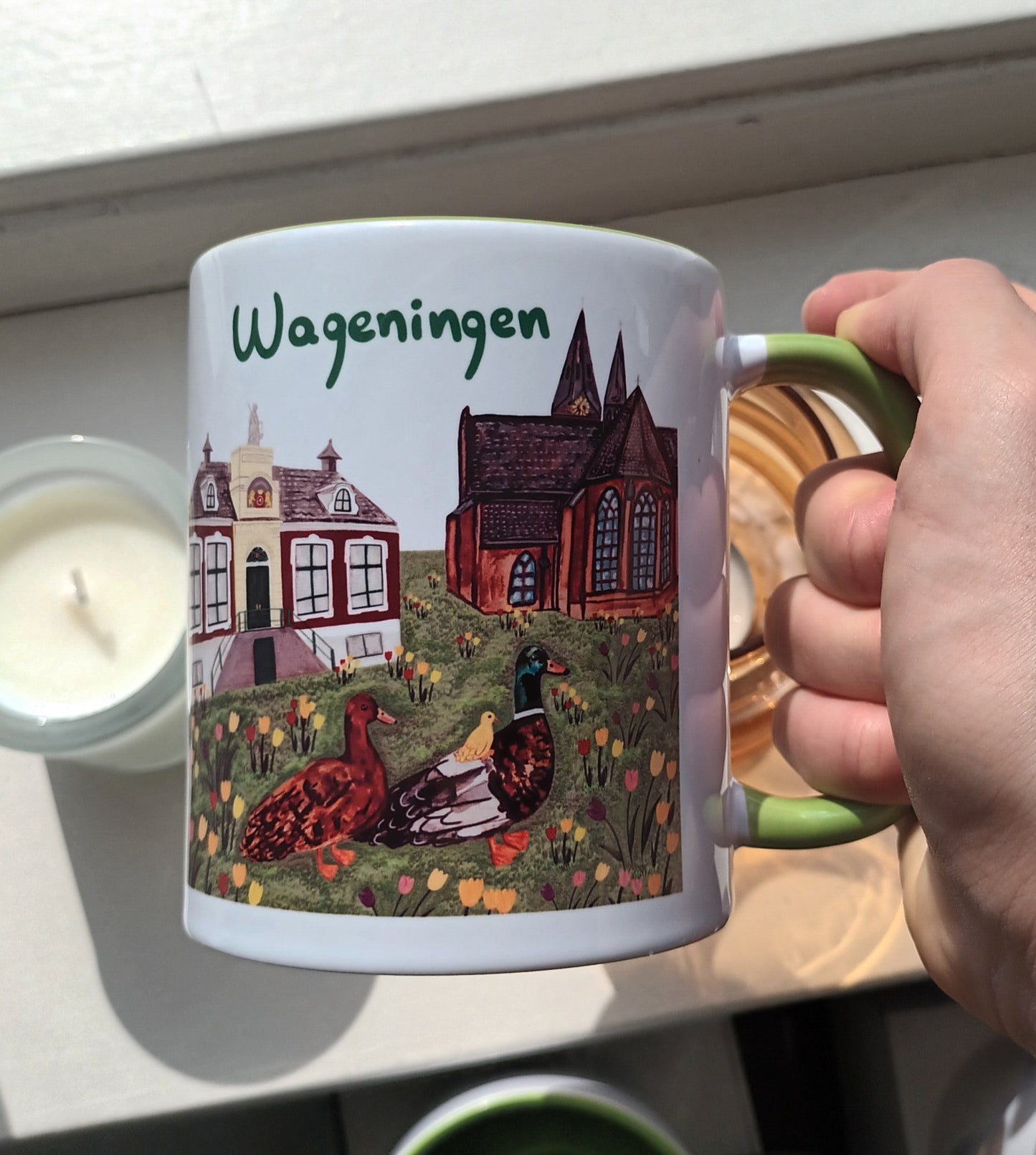 Mug with 'Wageningen' design held by a hand, next to a candle.