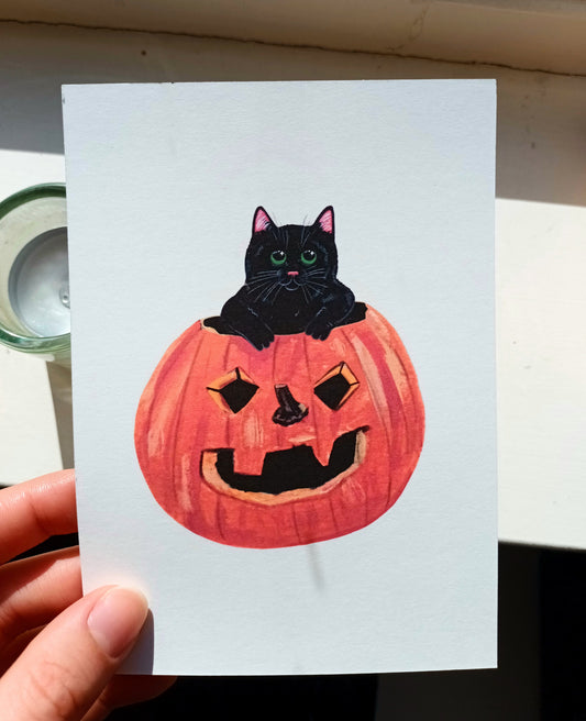 Card with a black cat in a pumpkin, held by a hand.