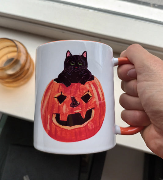 Mug with a Halloween design of a black cat in a jack-o'-lantern, held by a hand.
