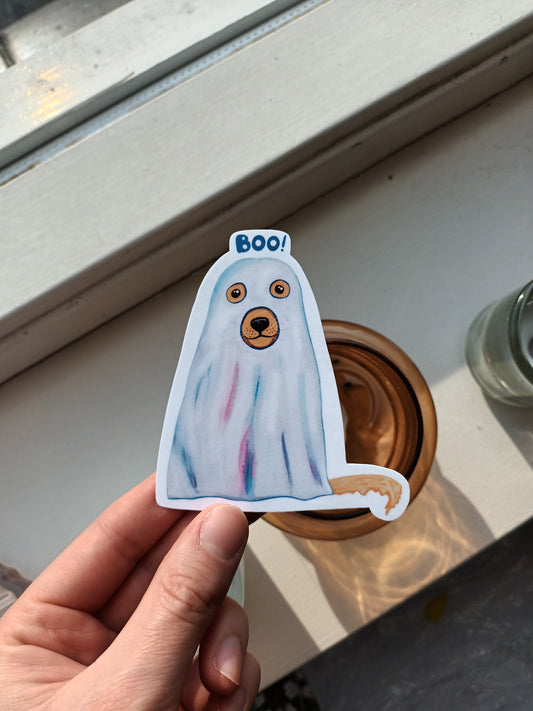 Hand holding a colourful dog ghost sticker with 'BOO!' text on a light surface.