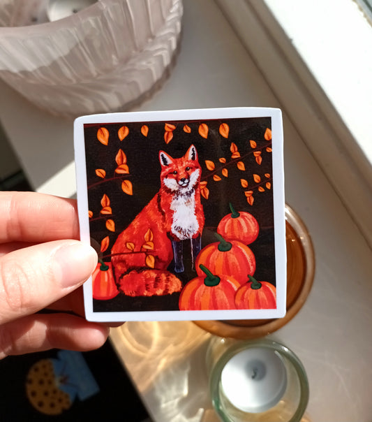 Sticker with a fox and pumpkins design held by a hand on a light surface.