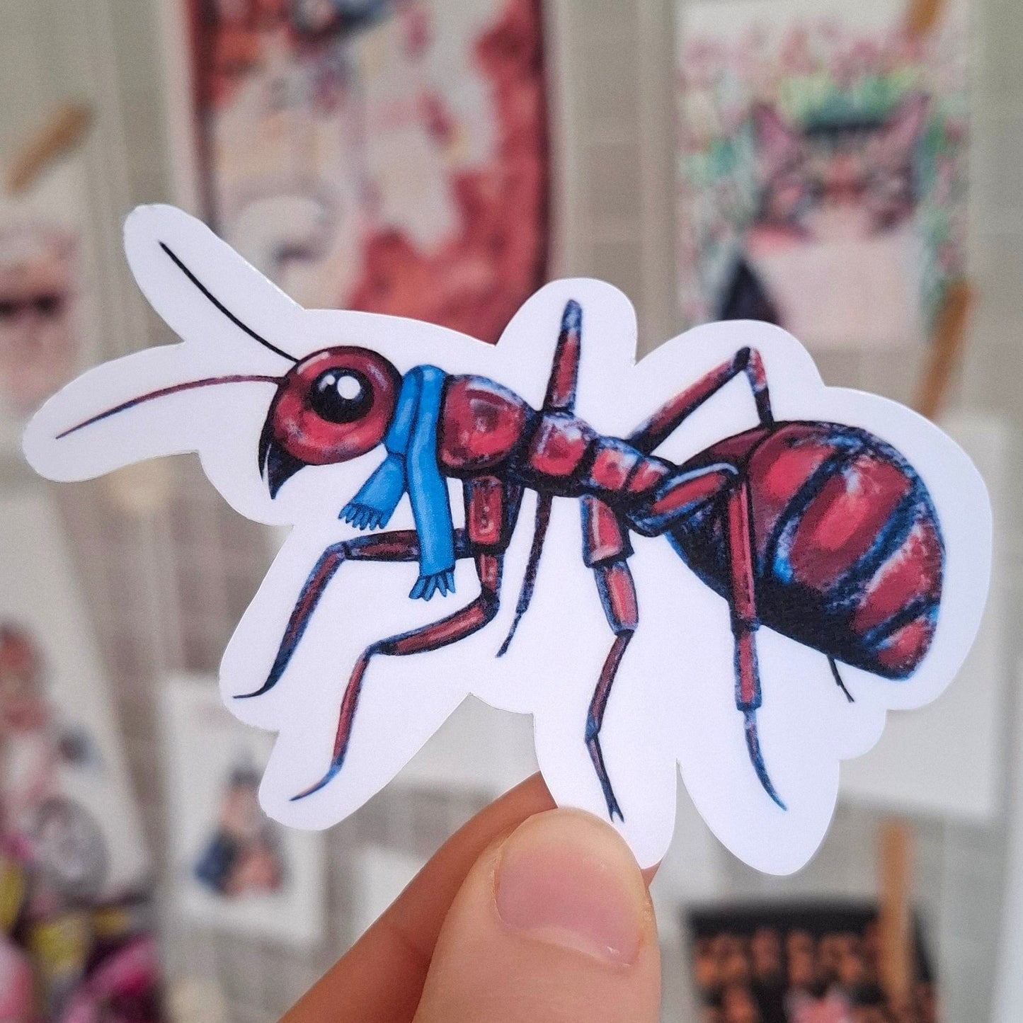 Hand holding a sticker of a red ant with a blue scarf, cute winter sticker.