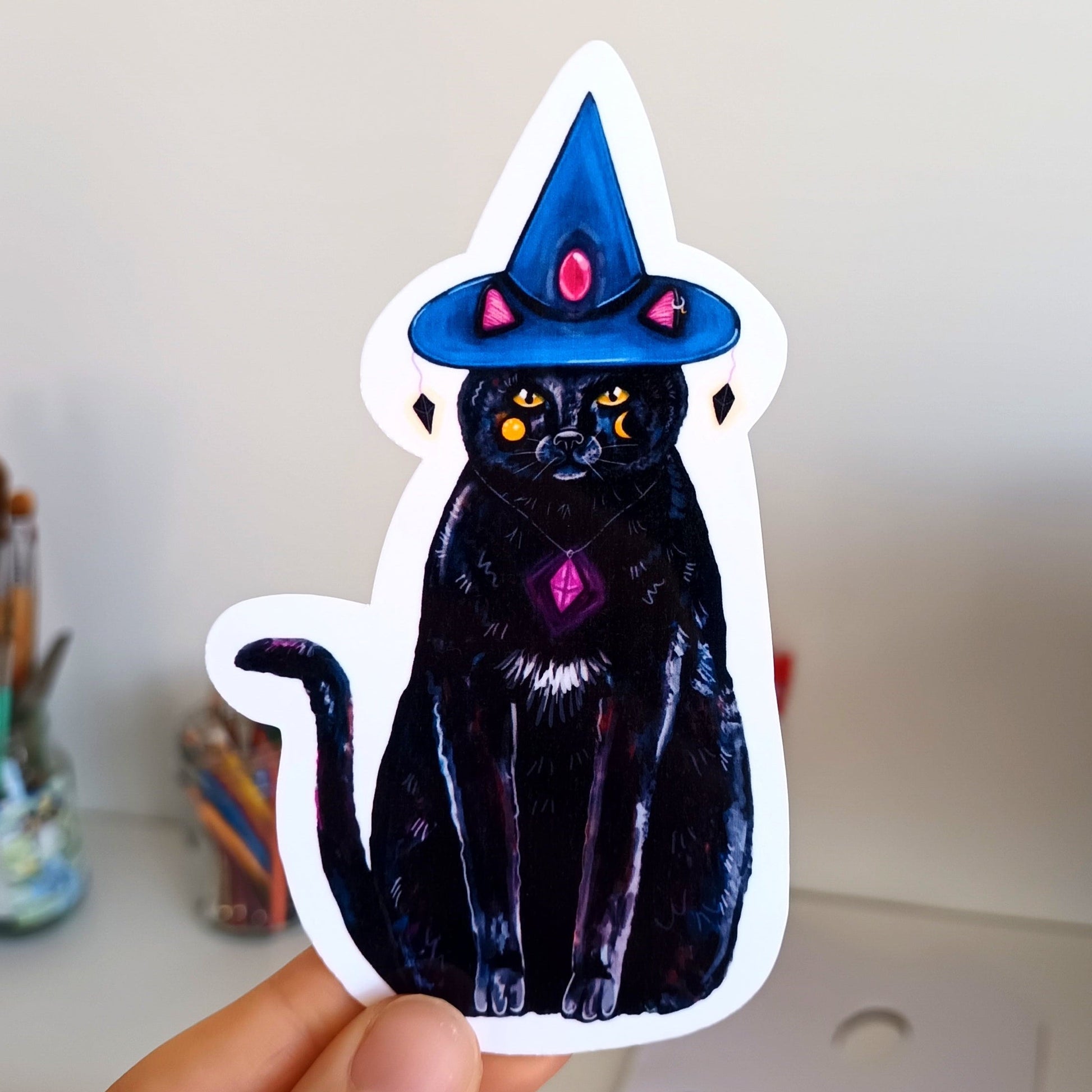 Sticker of a black cat with a wizard hat held by a hand on a white background.
