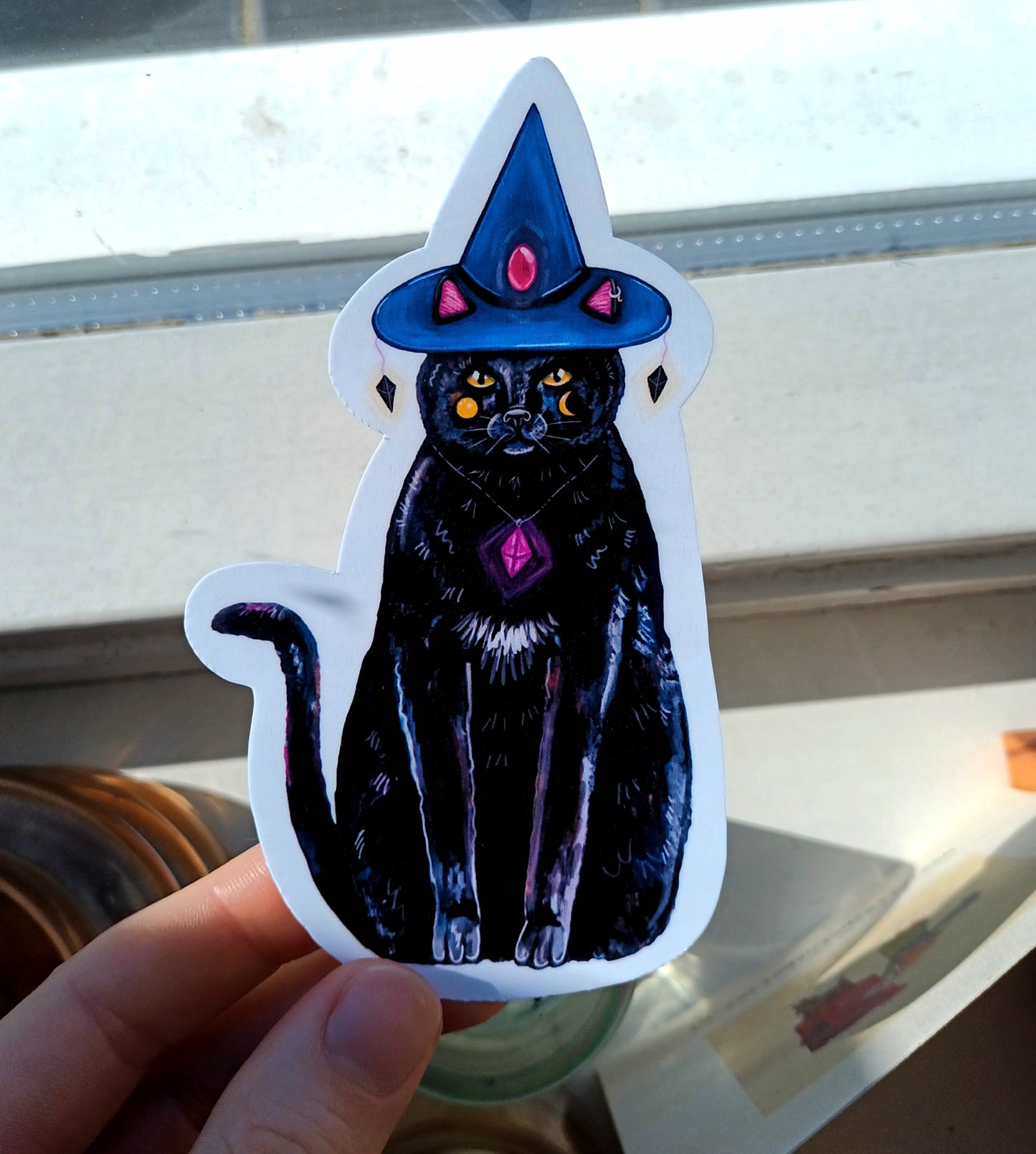 Sticker of a black cat wearing a witch hat held by a hand.