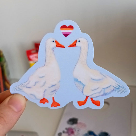 Hand holding a sticker of two gay geese with a heart between them