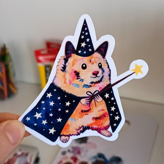 Hand holding a sticker of a hamster wizard with a wizard hat and robe.
