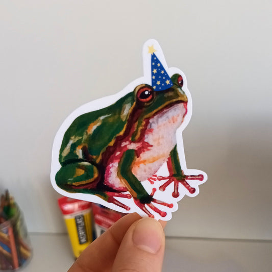 Hand holding a sticker of a frog wearing a wizard hat.
