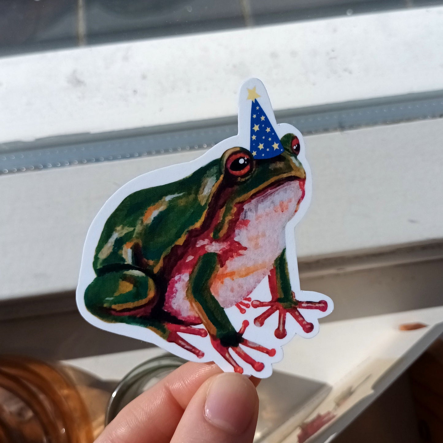 Hand holding a colourful frog sticker with a wizard hat.