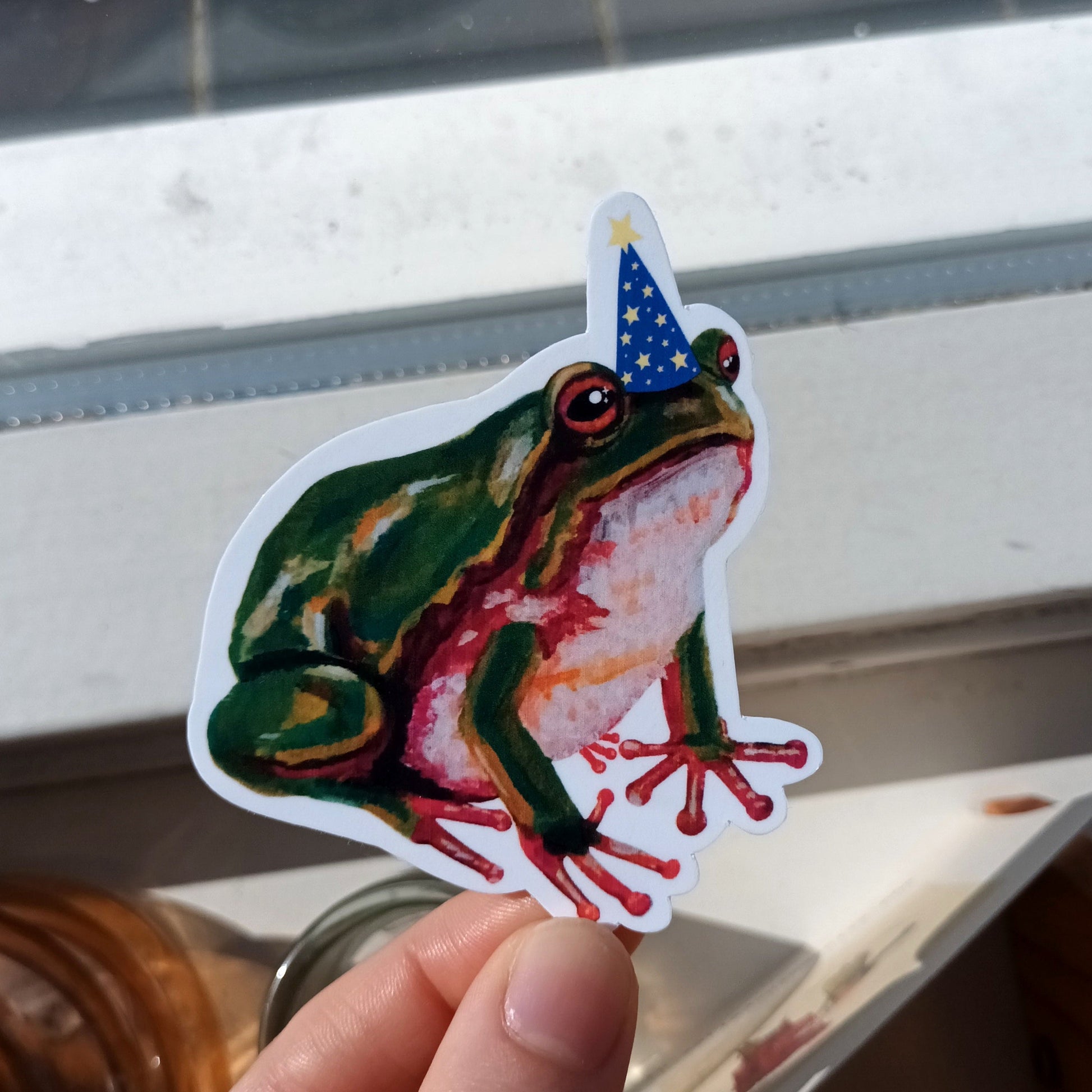 Hand holding a colourful frog sticker with a wizard hat.