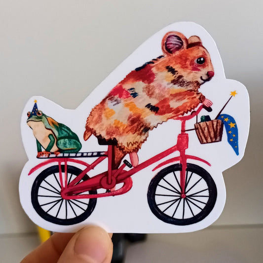 Hand holding a sticker of a hamster on a bicycle with a frog on the back.