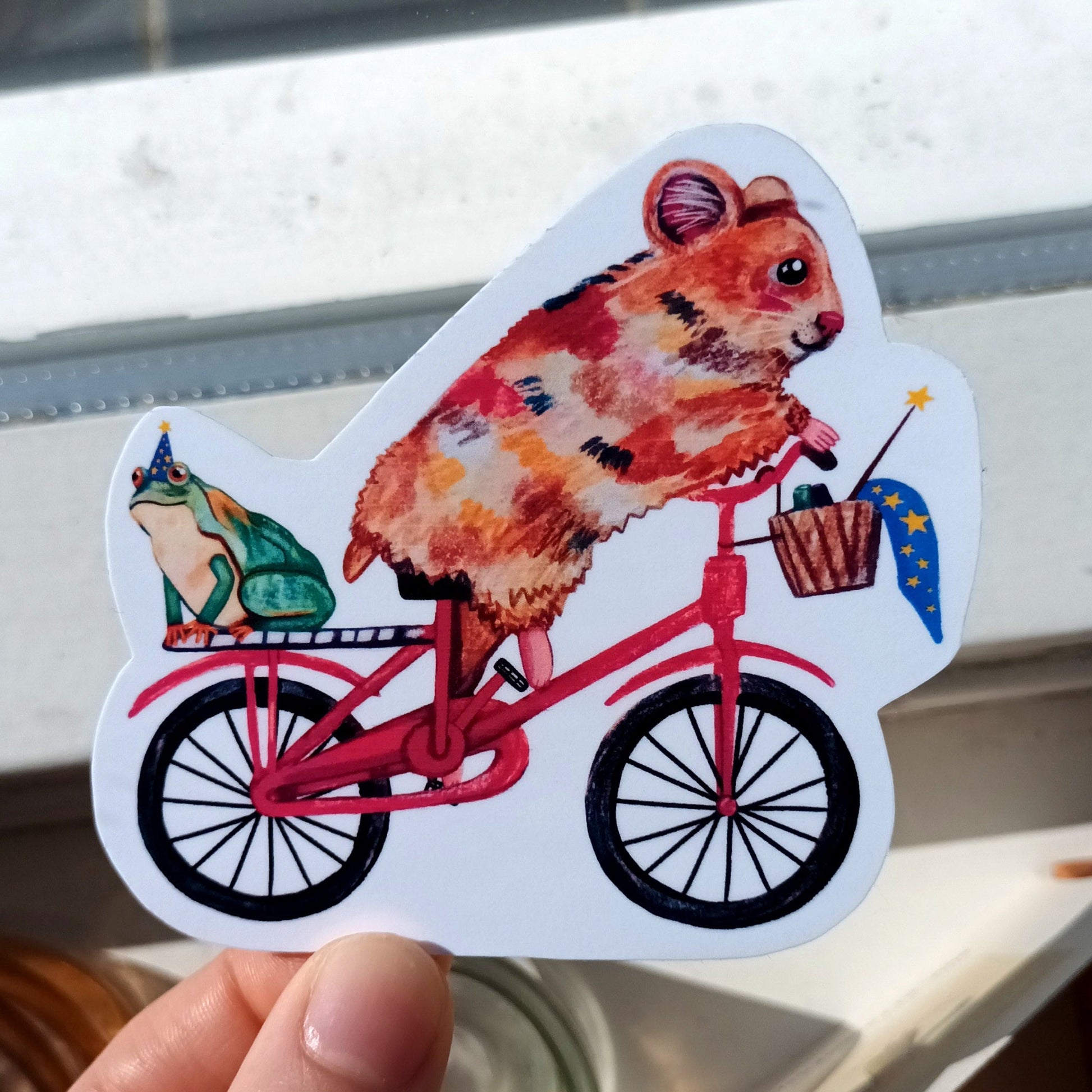 Hand holding a sticker of a hamster riding a bicycle with a basket.