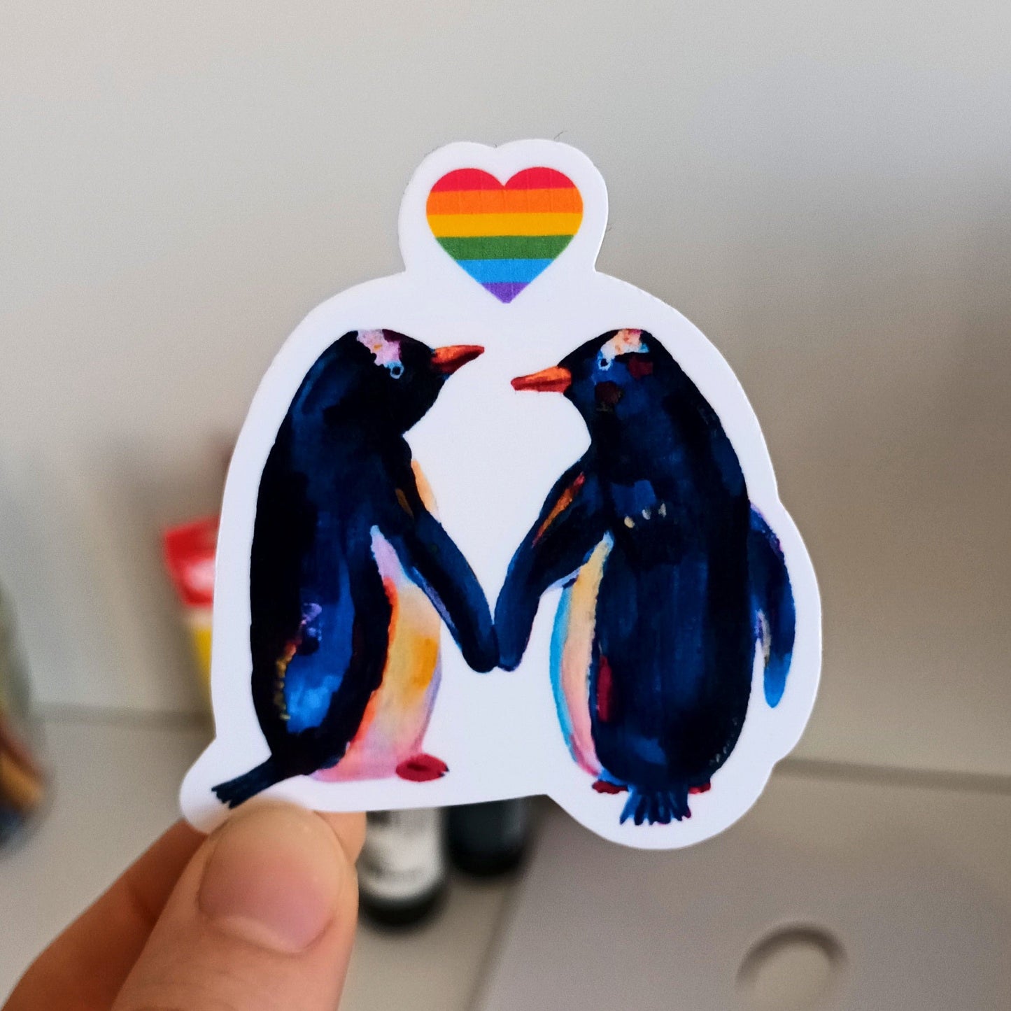 Hand holding a sticker of two penguins with a rainbow heart.