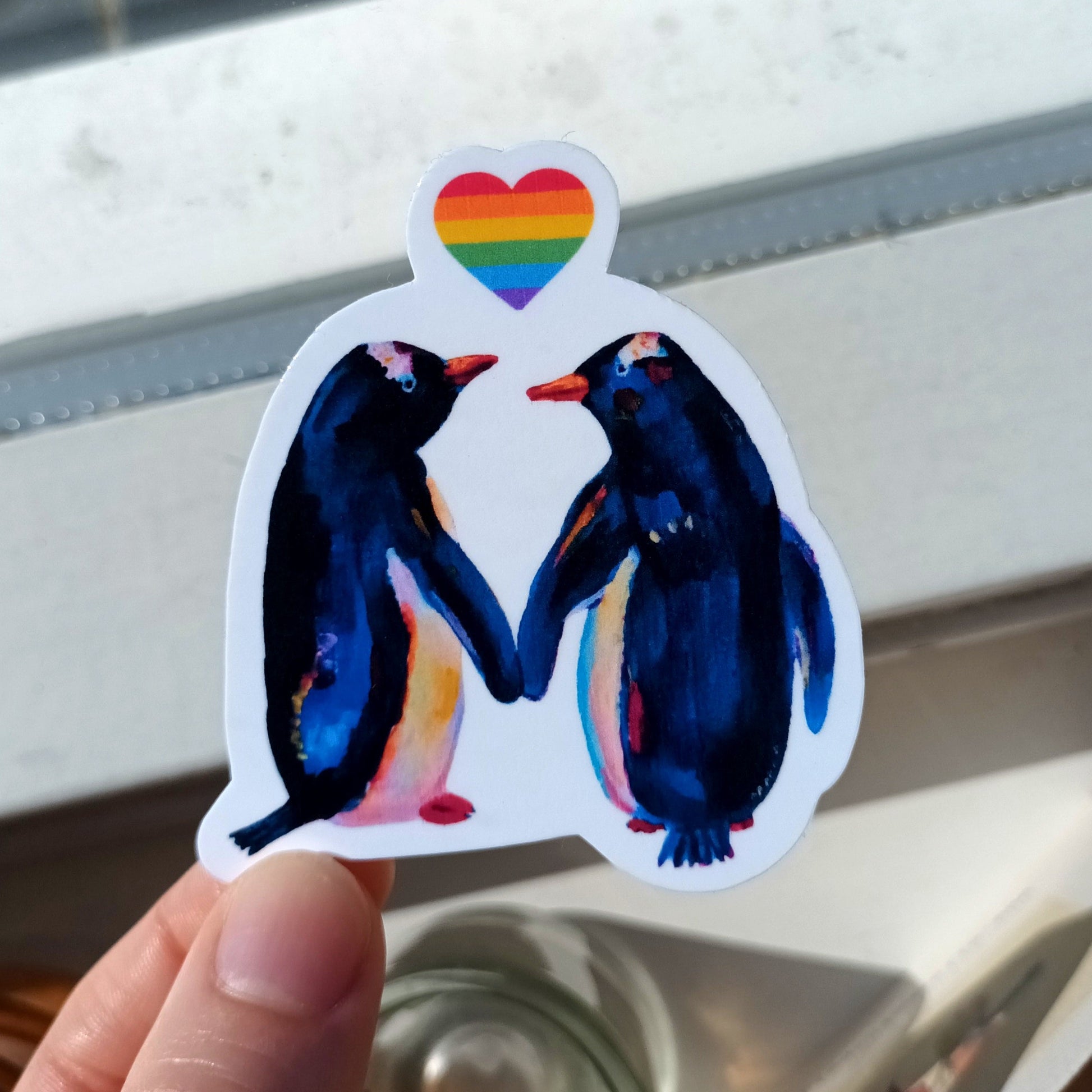 Sticker of two penguins holding hands with a rainbow heart above, held by a hand.