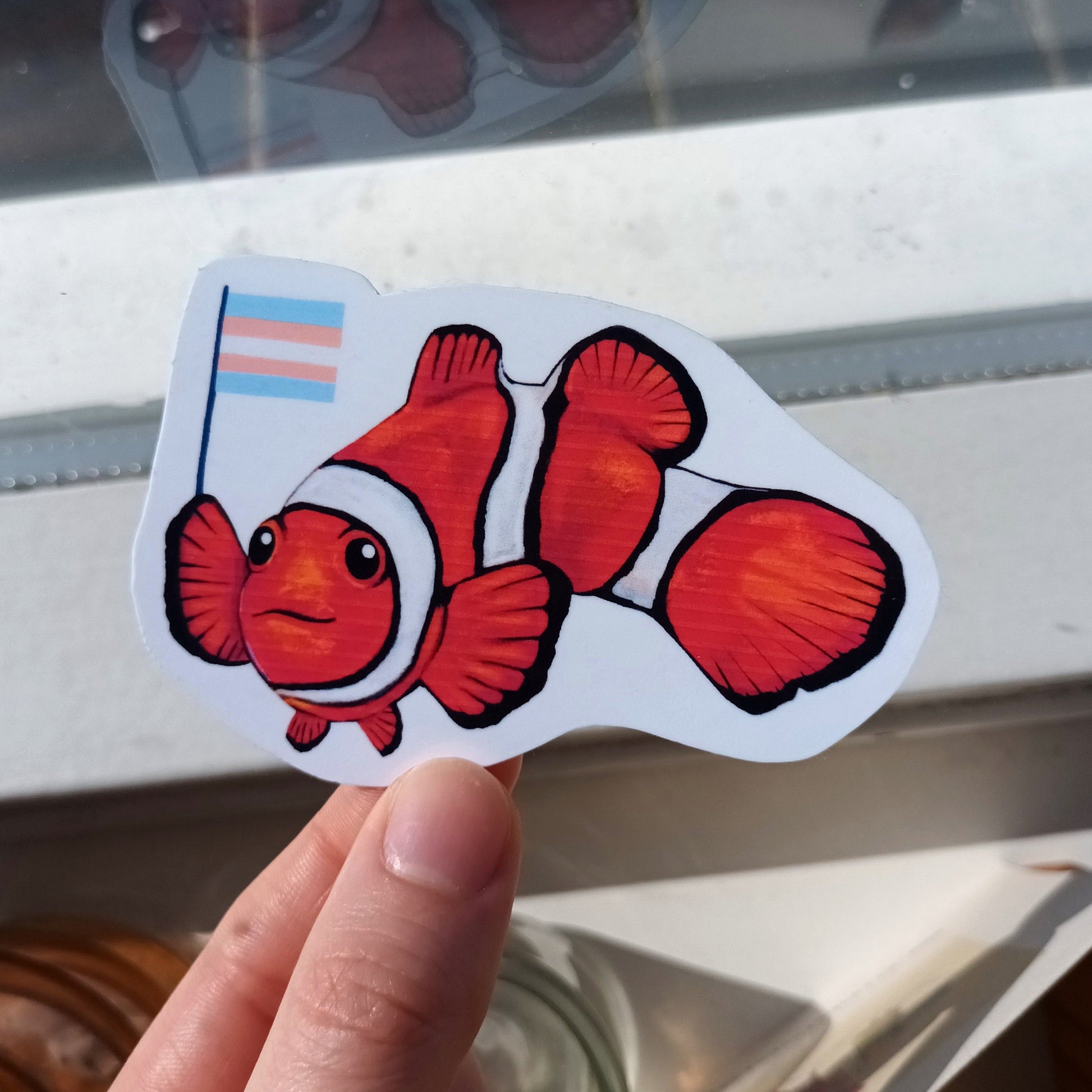 Hand holding a sticker of a clownfish with a small trans flag .