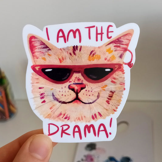 Sticker of a cat with sunglasses and 'I am the drama!' text held by a hand.