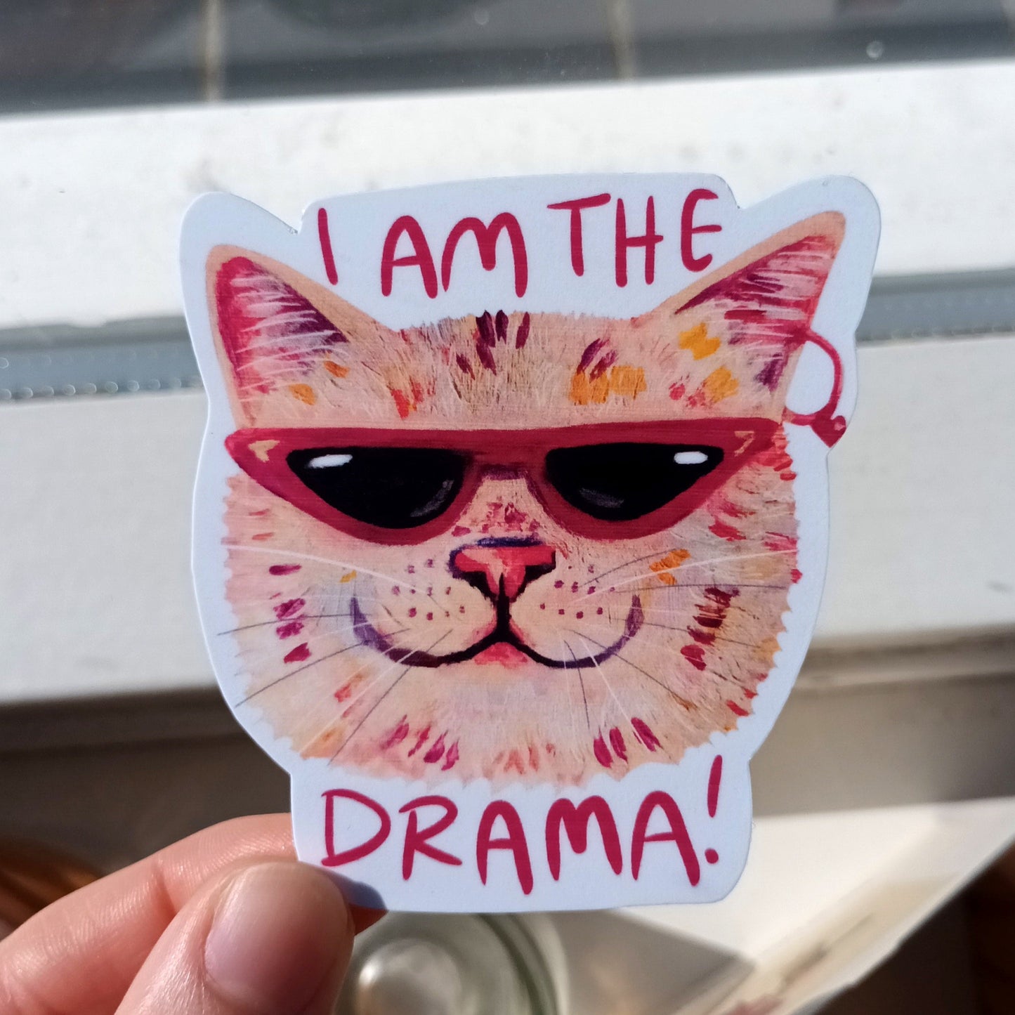 Sticker of a cat with sunglasses and 'I am the drama!' text held by a hand.