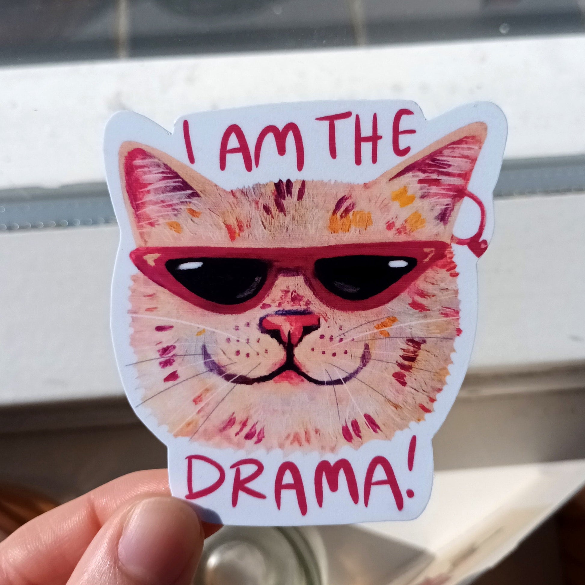 Sticker of a cat with sunglasses and 'I am the drama!' text held by a hand.