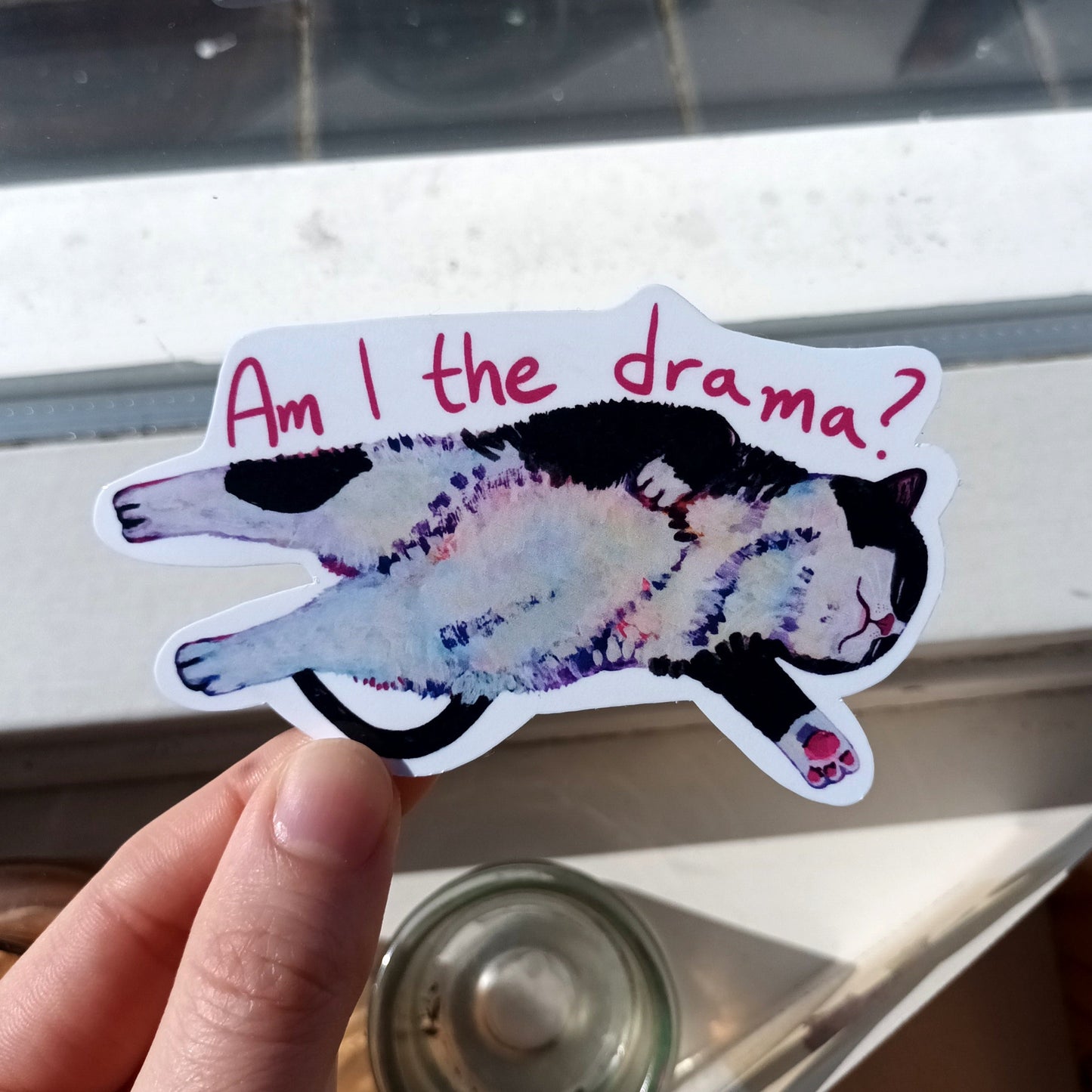 Sticker with a cat illustration and text 'Am I the drama?' held by a hand.