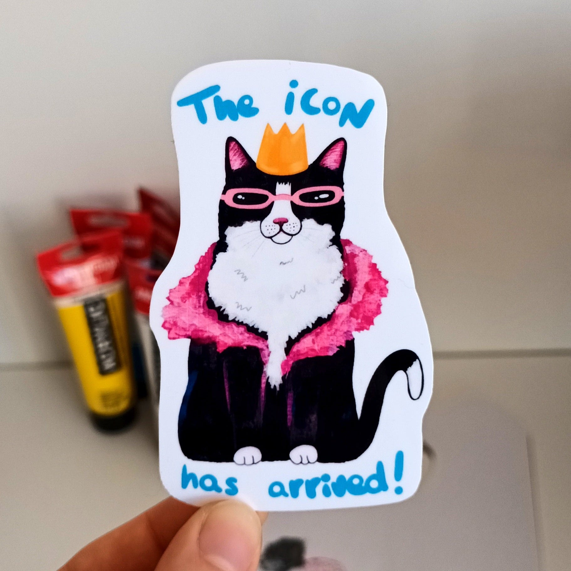Hand holding a sticker with a cat illustration and text 'The icon has arrived!' .