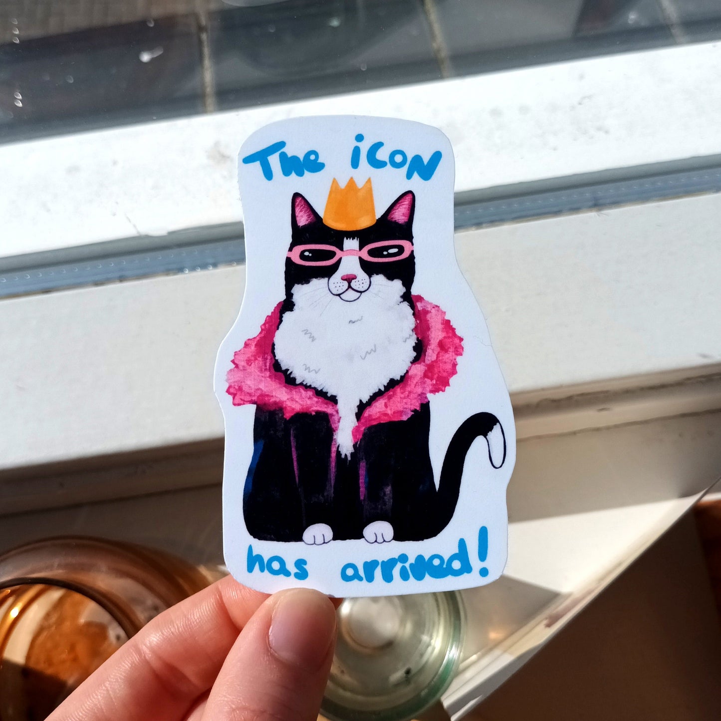 Sticker of a cat with sunglasses and a crown held by a hand, with text 'The icon has arrived!' .