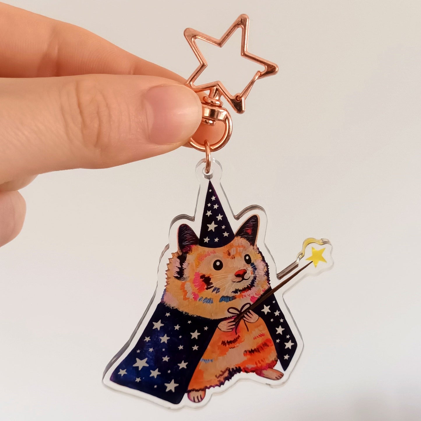 Hand holding a decorative keychain with a hamster in a witch hat and star design against a white wall.