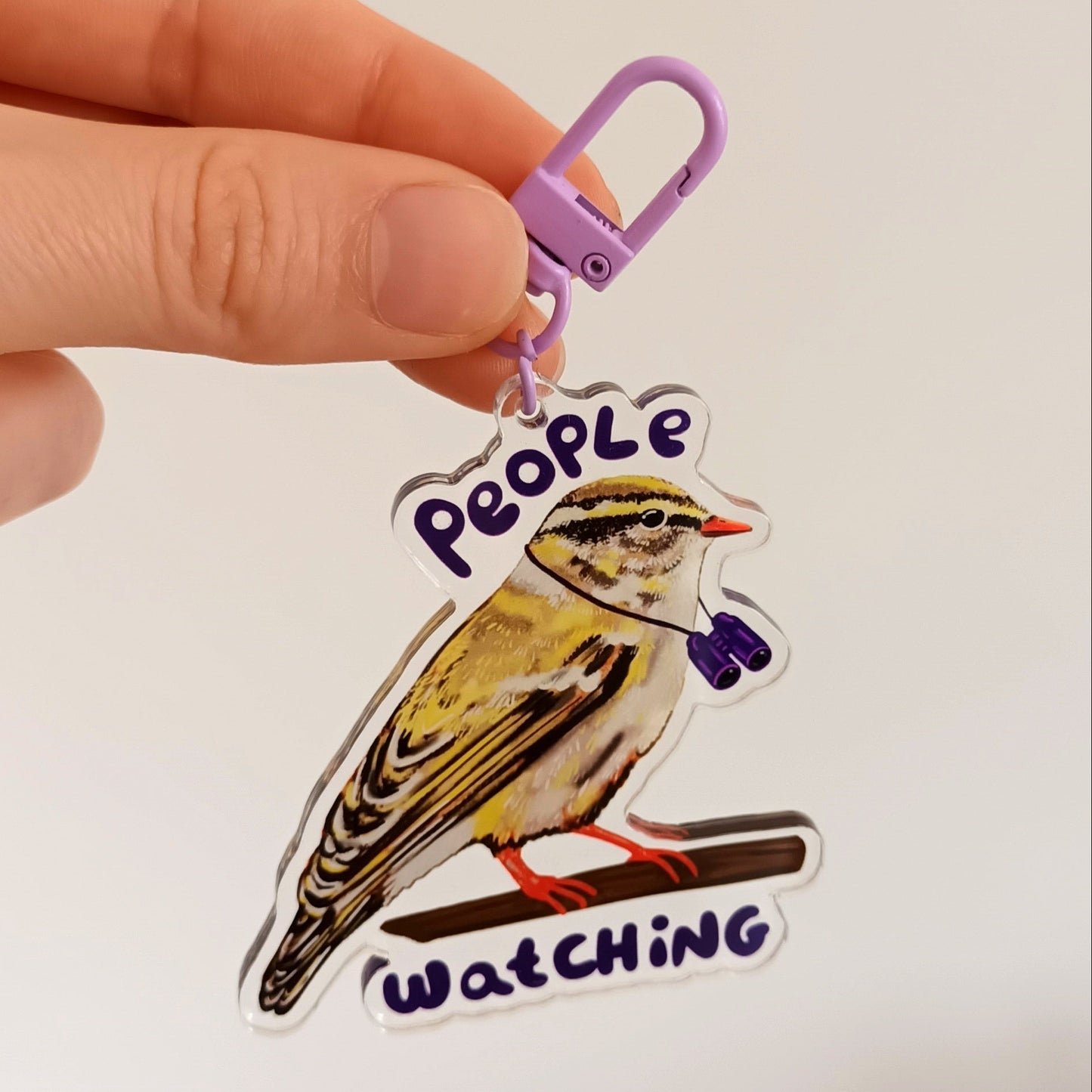 Hand holding a keychain with a bird design and 'People Watching' text, with art supplies in the background.