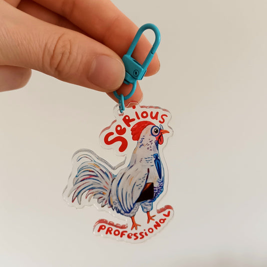 Hand holding a keychain with a rooster design and 'Serious Professional' text against a blurred background.