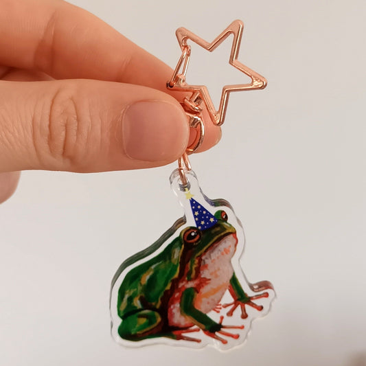 Hand holding a colorful frog-shaped keychain with starabin in an art studio setting.
